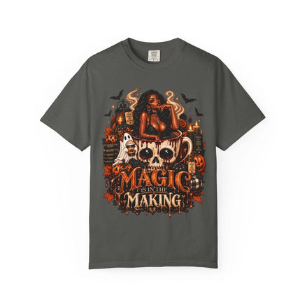 Unisex Garment-Dyed T-shirt in dark gray with a Halloween-themed graphic featuring a stylish woman, skull coffee mug, pumpkins, ghosts, bats, and candles with the text Magic Is in the Making